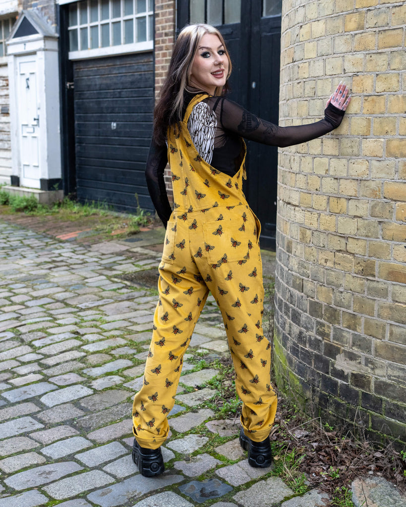 The model turns to reveal the back of Run & Fly’s Gold Bee Corduroy DungaBee Dungarees, showing the relaxed fit and adjustable straps. The all-over bee print contrasts against their black mesh top, with tattooed arms visible. They lean against a rustic brick wall on a cobbled street.