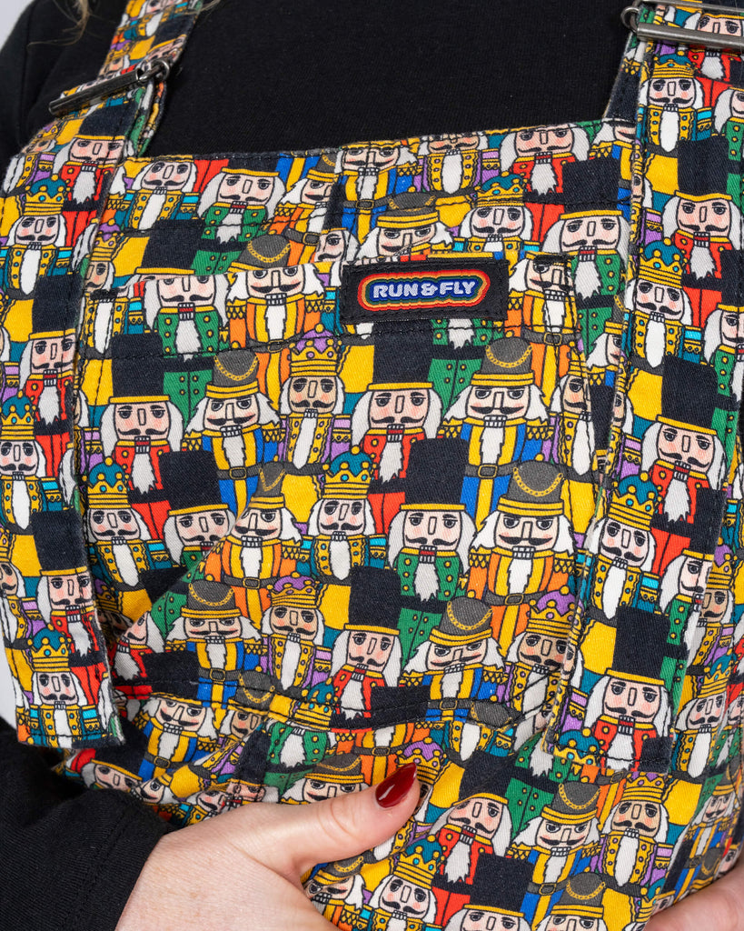 A medium close-up of Karina wearing the Nutcracker print dungarees, focusing on the chest panel. The bold Run & Fly logo is visible against the playful, colourful nutcracker pattern.