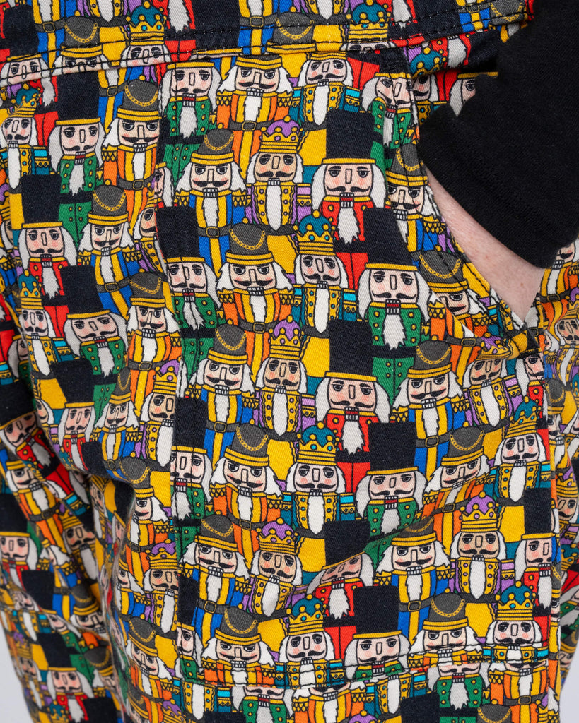 A detailed shot of the Nutcracker dungarees’ fabric. The vibrant characters, including kings and soldiers, are depicted in cheerful, festive colours. Karina showcases the front pocket design with her hand resting inside.
