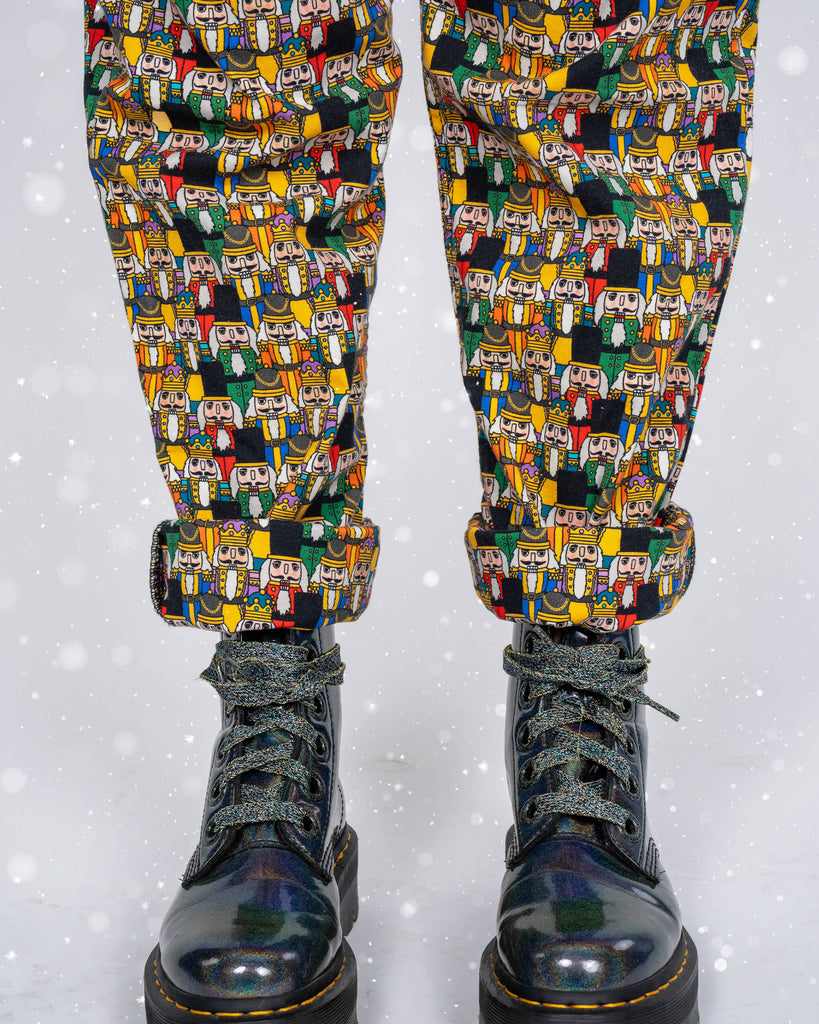 A close-up of Nutcracker print dungarees by Run & Fly. The pattern features colourful nutcracker characters in red, green, yellow, and black on a dark background, paired with shiny lace-up boots. Model Karina’s hand is in the pocket, showing off the design details.