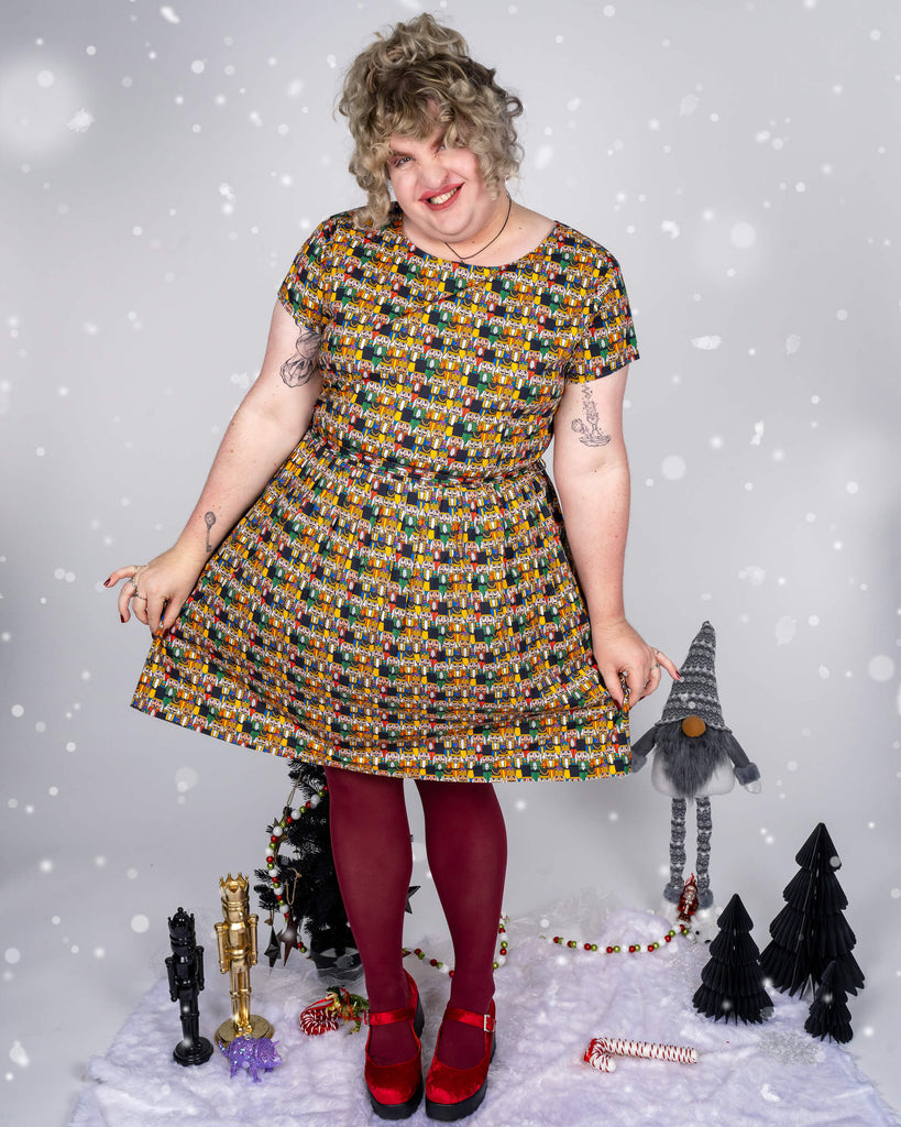 Full-length shot of Maddie wearing a festive dress by Run & Fly, featuring a colourful Nutcracker print designed by Waffle Mama Design. Maddie pairs the dress with maroon tights and red shoes, standing against a snowy backdrop with holiday decorations, including miniature trees, a gnome, and nutcracker figures.