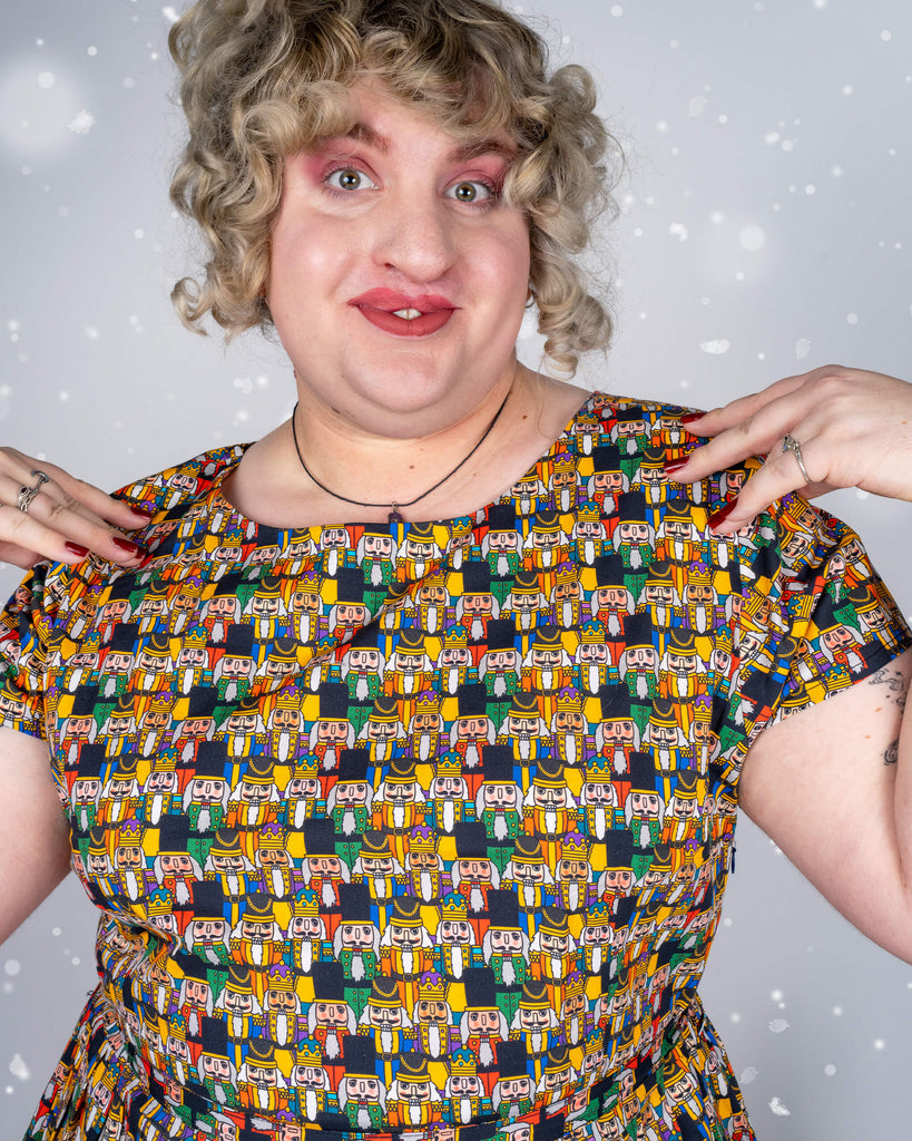 Close-up of Maddie’s expression, showing excitement with hands near the collar of the Nutcracker print dress. Maddie’s curly hair, bold makeup, and joyful look add a fun holiday vibe to the image, against a light, snowy background.