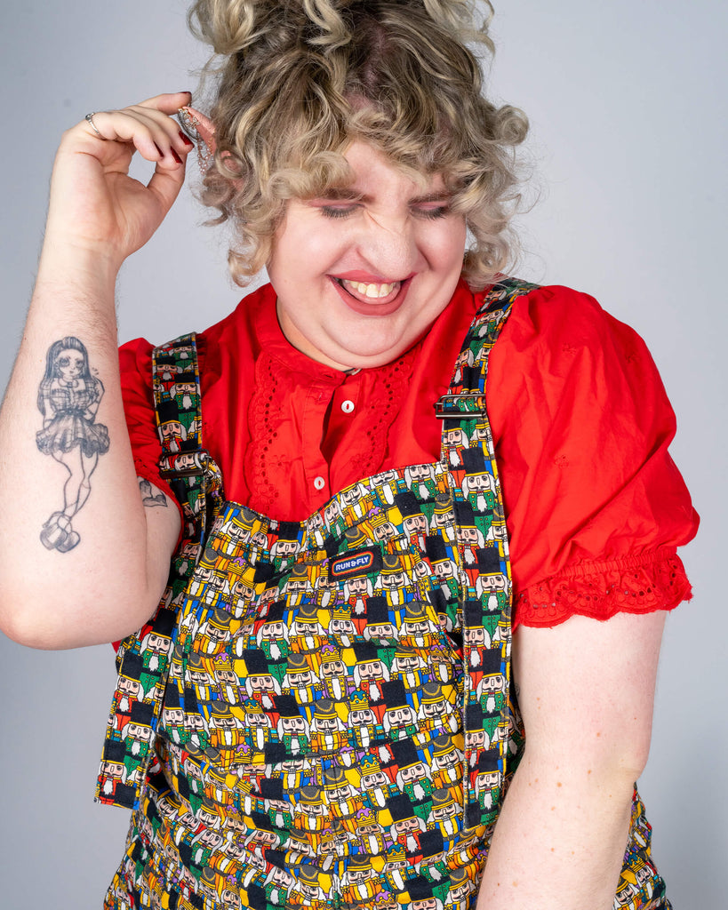 Model Maddie, dressed in Run & Fly’s Nutcracker print dungarees, laughs while holding an ear adorned with sparkly holiday accents. Her outfit includes a red blouse, maroon tights, and red velvet shoes, adding to the holiday charm. The scene includes seasonal decorations like a mini Christmas tree, enhancing the festive mood.
