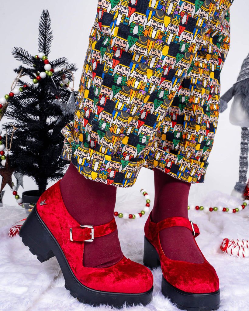A close-up of Maddie’s feet in red velvet shoes and maroon tights, highlighting the intricate Nutcracker print on the Run & Fly dungarees. The festive details around, including a small decorated tree, add a holiday touch to the scene.