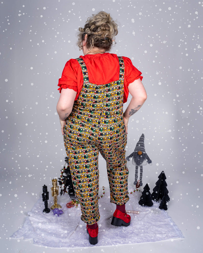 Model Maddie stands with her back to the camera in Run & Fly’s Nutcracker print dungarees, showcasing the vibrant nutcracker design. Paired with a red blouse and surrounded by holiday decorations, the image captures a festive, playful vibe.