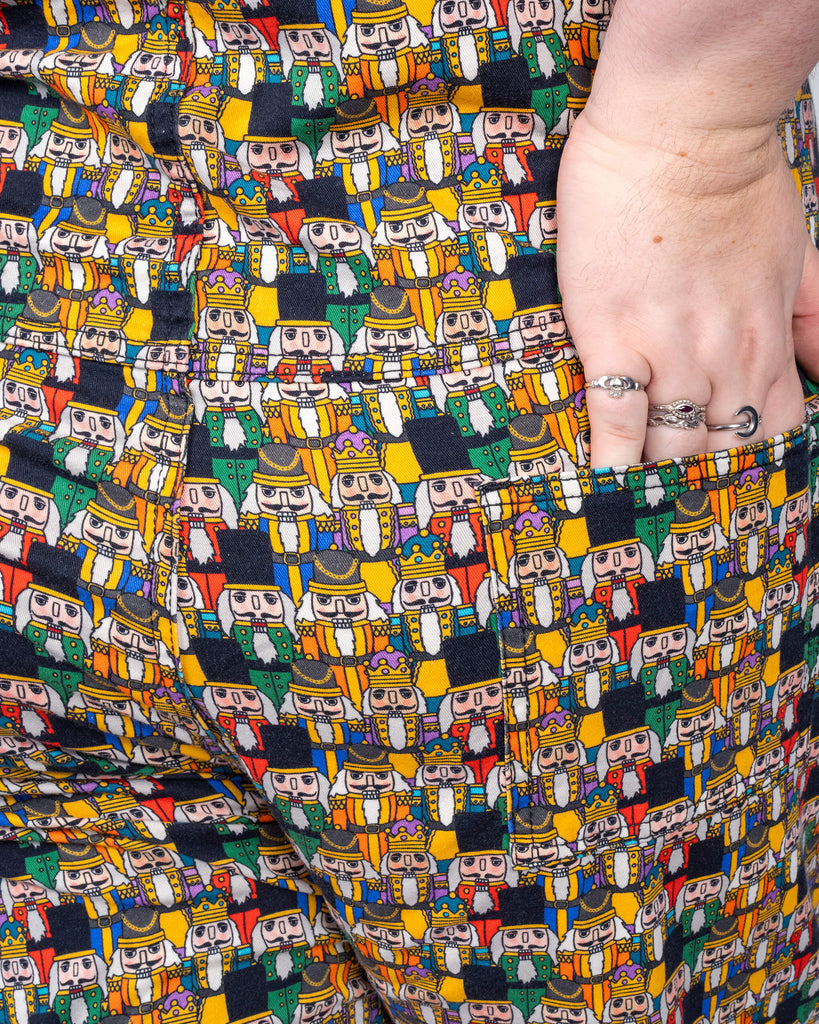 Maddie shows off the Nutcracker print dungarees by Run & Fly, featuring a colourful array of nutcracker characters. Her hand casually rests in the back pocket, displaying the detailed, lively design. The festive scene in the background enhances the holiday spirit.