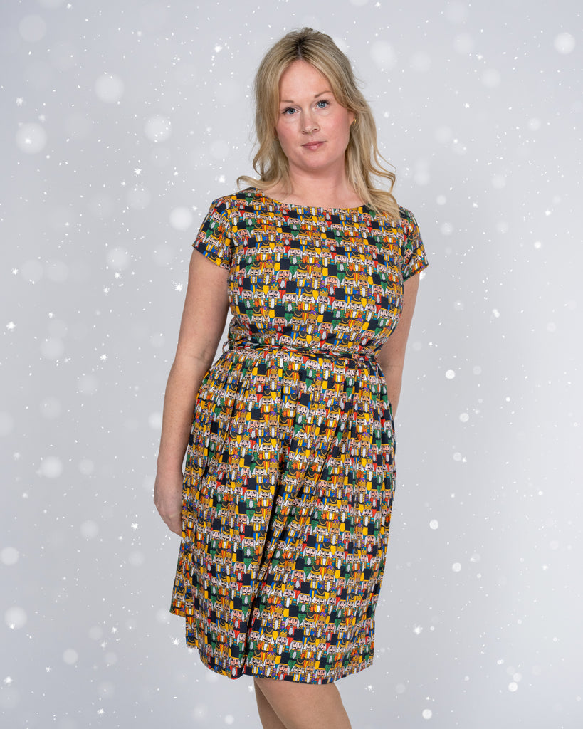  full-length view of the Nutcracker print dress, highlighting its knee-length A-line skirt and tie waist. The model stands with a confident pose in front of a snow-flecked background, wearing black lace-up boots.