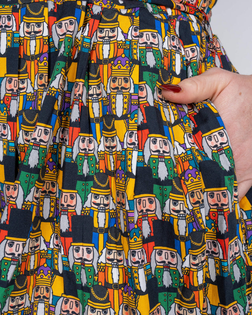 A close-up of the dress’s pleated skirt with hands in the pockets, showing off the intricate Nutcracker designs. The model’s nails are painted deep red, complementing the festive theme.