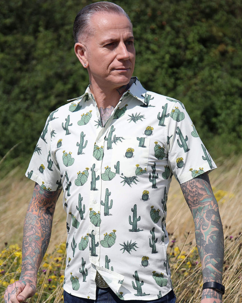 A heavily tattooed model is stood outside in a tall grass field wearing the cactus short sleeve shirt with denim jeans. He is facing the camera and posing with both hands out at his sides and looking off into the right. Photo is cropped from the knees up.