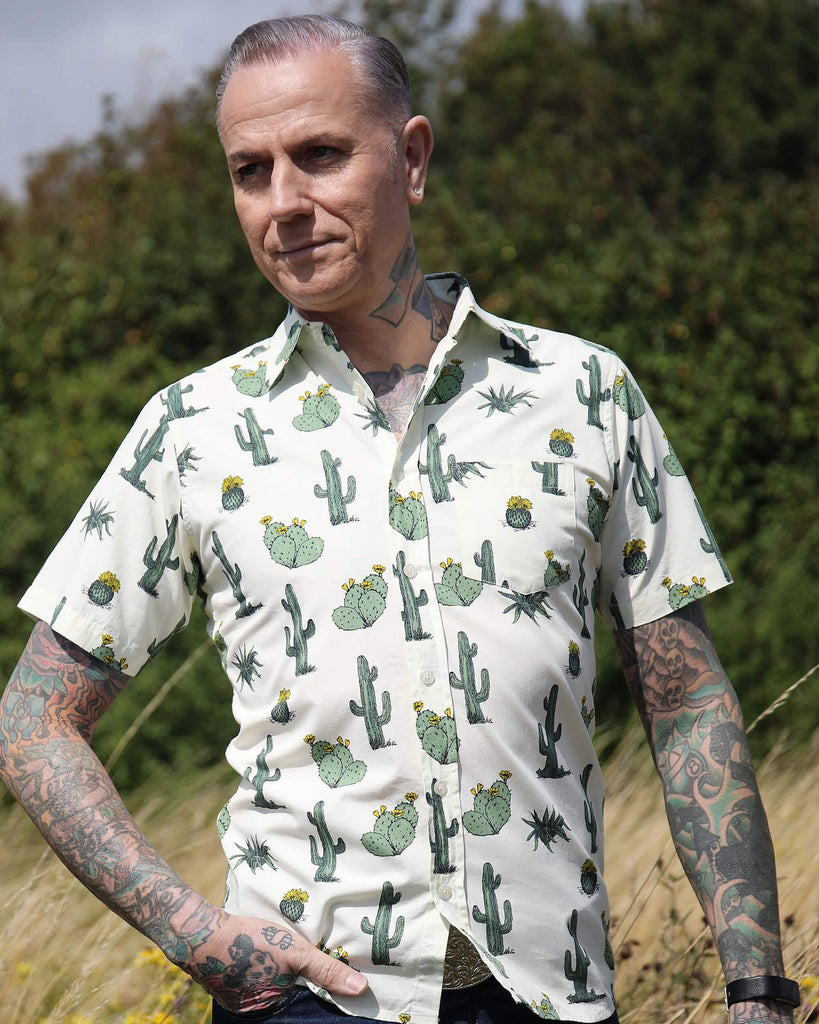 A heavily tattooed model is stood outside in a tall grass field wearing the cactus short sleeve shirt with denim jeans. He is facing towards the camera with one hand in his front pocket with the other resting by his side, he is looking off to the left.