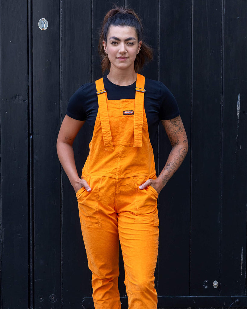 Model is wearing Highlighter Orange Stretch Corduroy Dungarees paired with a black t-shirt and black chunky boots. The dungarees are a bright orange with Run&Fly logo on the front pocket on chest. Model has her hands in front pockets. The bottoms of the legs are turned up.