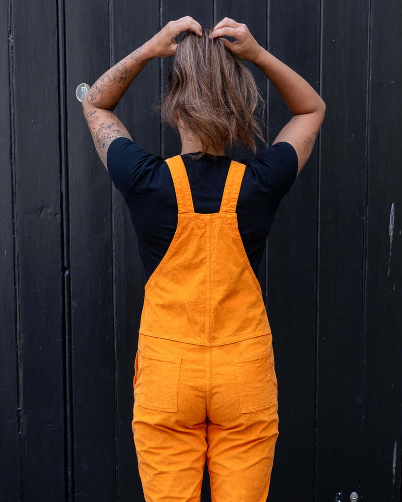 Model is wearing Highlighter Orange Stretch Corduroy Dungarees paired with a black t-shirt and black chunky boots. The dungarees are a bright orange colour. Model has her back to the camera and is running her hands through her hair. There are two pockets on the bum of the dungarees and bottoms of the legs are turned up.