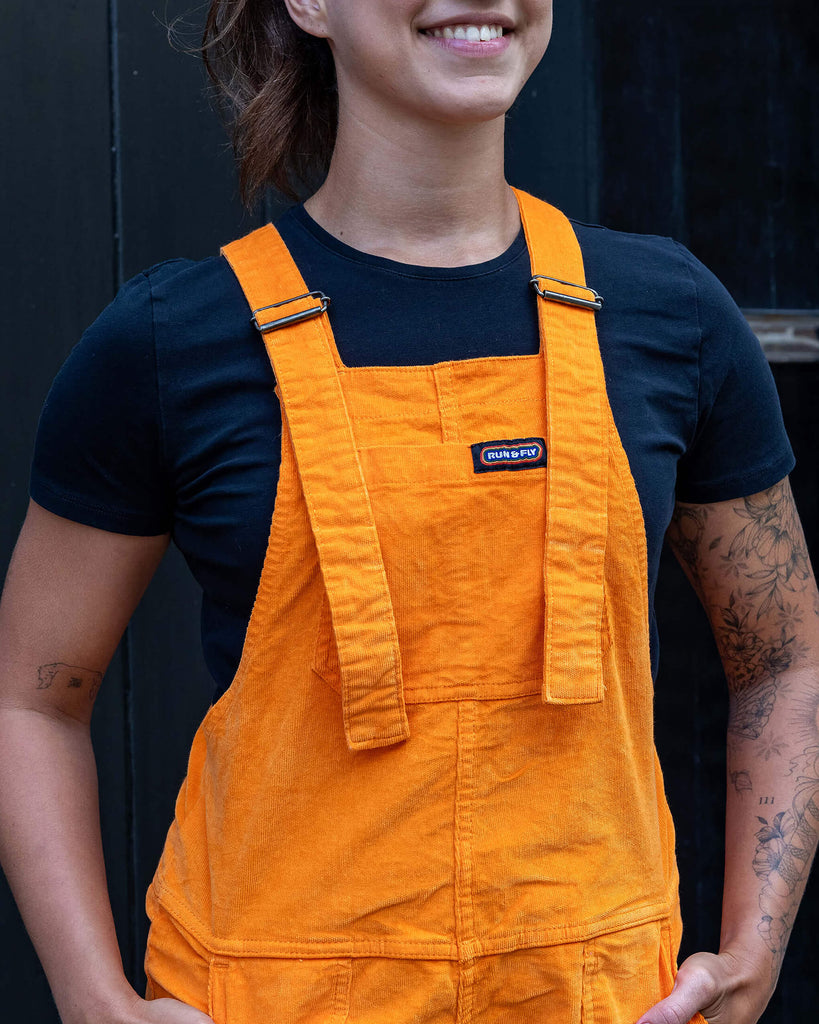 Model is wearing Highlighter Orange Stretch Corduroy Dungarees paired with a black t-shirt. The dungarees are a bright orange with Run&Fly logo on the front pocket on chest. Model is smiling and has her hands in front pockets.