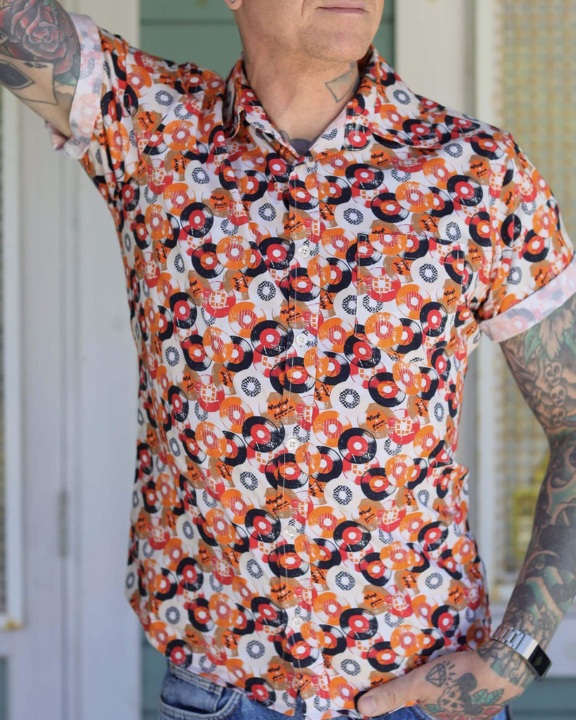 Jim is stood outside in a doorway wearing the orange record short sleeve shirt with blue denim jeans. He is heavily tattooed and posing with one arm leaning up against the doorway with the other hand in his front jean pocket. Photo is cropped from the chin to hips.