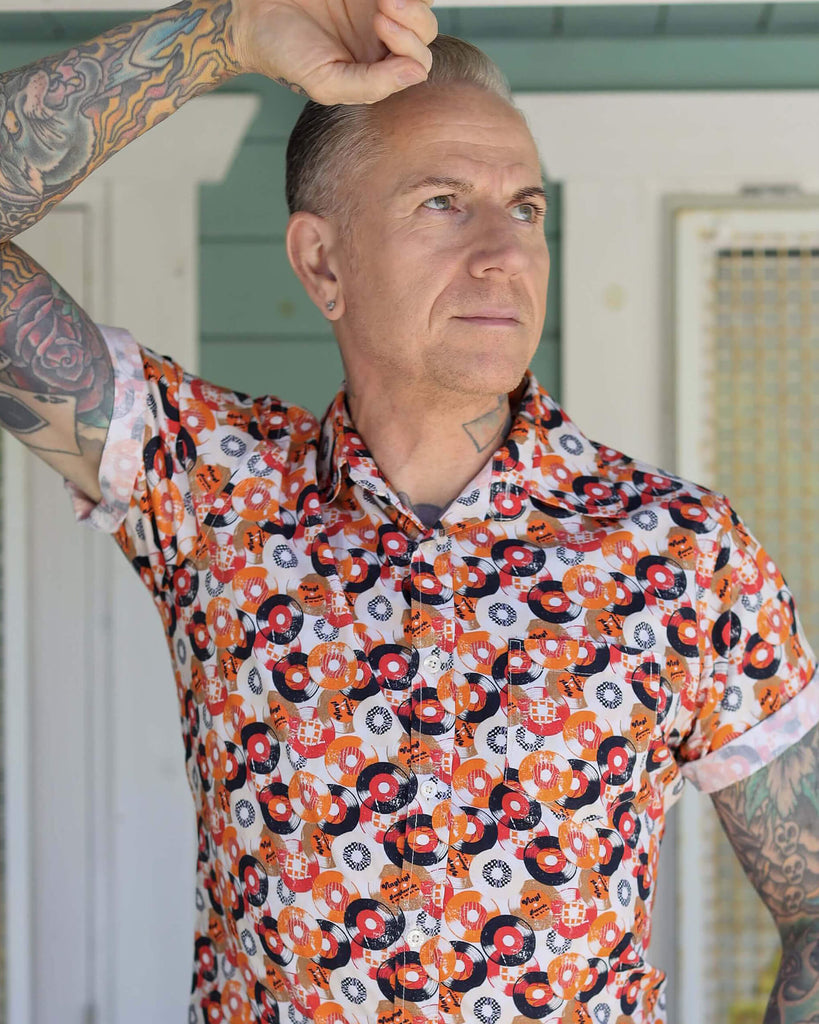 Jim is stood outside in a doorway wearing the orange record short sleeve shirt with blue denim jeans. He is heavily tattooed and posing with one arm leaning up against the doorway with the other hand in his front jean pocket. Photo is cropped from the waist up.