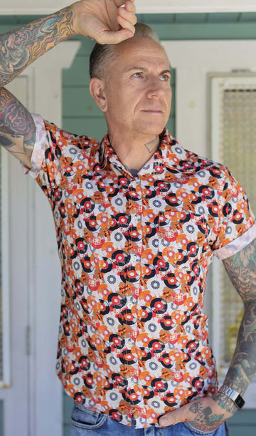 Jim is stood outside in a doorway wearing the orange record short sleeve shirt with blue denim jeans. He is heavily tattooed and posing with one arm leaning up against the doorway with the other hand in his front jean pocket. Photo is cropped from the hips up.
