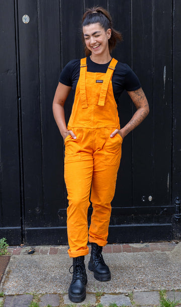 Model is wearing Highlighter Orange Stretch Corduroy Dungarees paired with a black t-shirt and black chunky boots. The dungarees are a bright orange with Run&Fly logo on the front pocket on chest. Model is smiling and has her hands in front pockets. The bottoms of the legs are turned up.