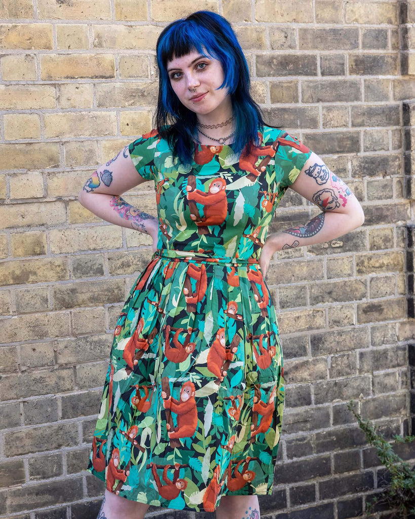 Faeryn is stood outside in Hove in a mews wearing Orangutan Stretch Belted Tea Dress with Pockets paired with black boots. The green dress has an all over orangutan print with leaves and branches. Faeryn is stood facing the camera with their hands on their hips and Brian the dog is stood next to them. 