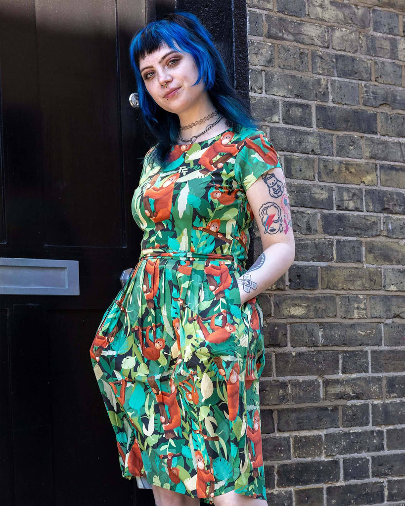 Faeryn is stood outside in Hove in a mews wearing Orangutan Stretch Belted Tea Dress with Pockets paired with black boots. The green dress has an all over orangutan print with leaves and branches. Faeryn is posing with their hand in the dress pocket. 