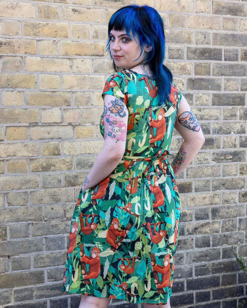 Faeryn is stood outside in Hove in a mews wearing Orangutan Stretch Belted Tea Dress with Pockets paired with black boots. The green dress has an all over orangutan print with leaves and branches. Faeryn is stood with their back to the camera as is looking back over their shoulder with their hands in the dress pockets. 