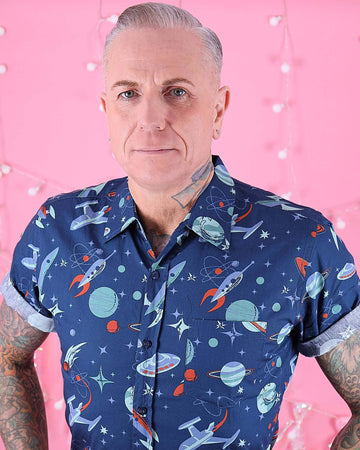 Jim is stood in front of a pink studio background wearing the outer space retro short sleeve shirt with brown corduroy trousers. He is heavily tattooed with silver swept back hair. He is posing facing the camera with both hands in his front trouser pockets. Photo is cropped from the hips up.