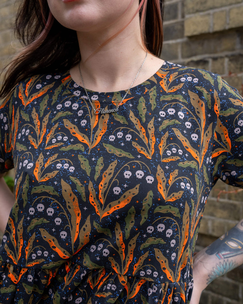 A front view of a person wearing a Run & Fly oversized smock dress. The spooky design features orange and green foliage, tiny skulls, and stars, perfect for a Halloween look. The person is wearing a necklace with the name "Florence" and has tattoos on their arms. The brand is a small ethical slow fashion company.