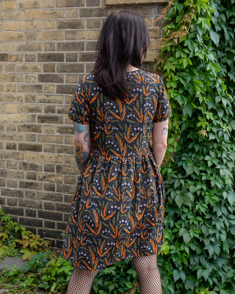 A person with tattoos stands with their back to the camera, wearing an oversized smock dress by Run & Fly. The dress features a Halloween-inspired print with orange and green leaves, tiny skulls, and stars. The person is standing against a brick wall and is wearing fishnet tights. The brand is a small ethical slow fashion company.