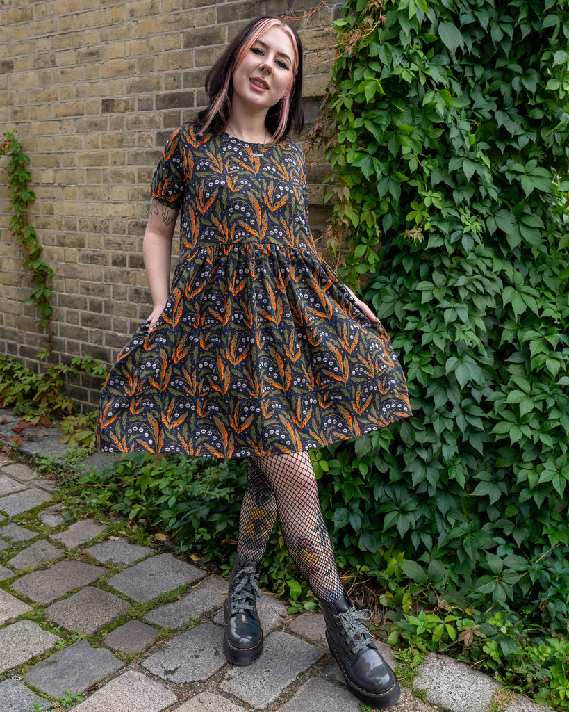 A person wearing an oversized smock dress from Run & Fly poses outdoors in front of a brick wall and greenery. The dress has an all-over print of skulls, orange and green leaves, and stars. The person completes the look with fishnet tights and black platform boots. The brand is a small ethical slow fashion company.