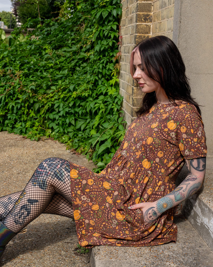 A person sits outdoors, leaning against a brick wall, wearing a Run & Fly smock dress with an autumn print of pumpkins and leaves. Fishnet tights and boots complete the look, with visible tattoos on their legs and arms. The brand is a small ethical slow fashion company.