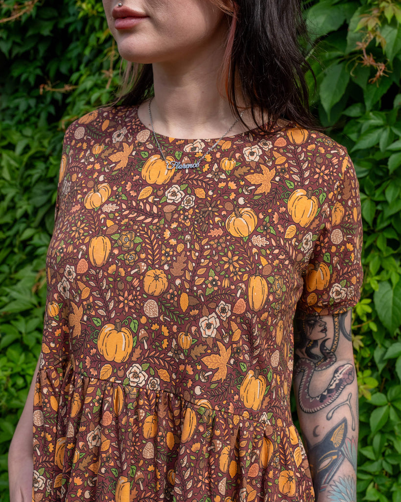 A close-up of a person wearing a Run & Fly smock dress, featuring a brown fabric decorated with orange pumpkins, mushrooms, and leaves. The person's arm tattoos are visible, and a "Florence" necklace is part of the outfit. The background is green foliage. The brand is a small ethical slow fashion company.