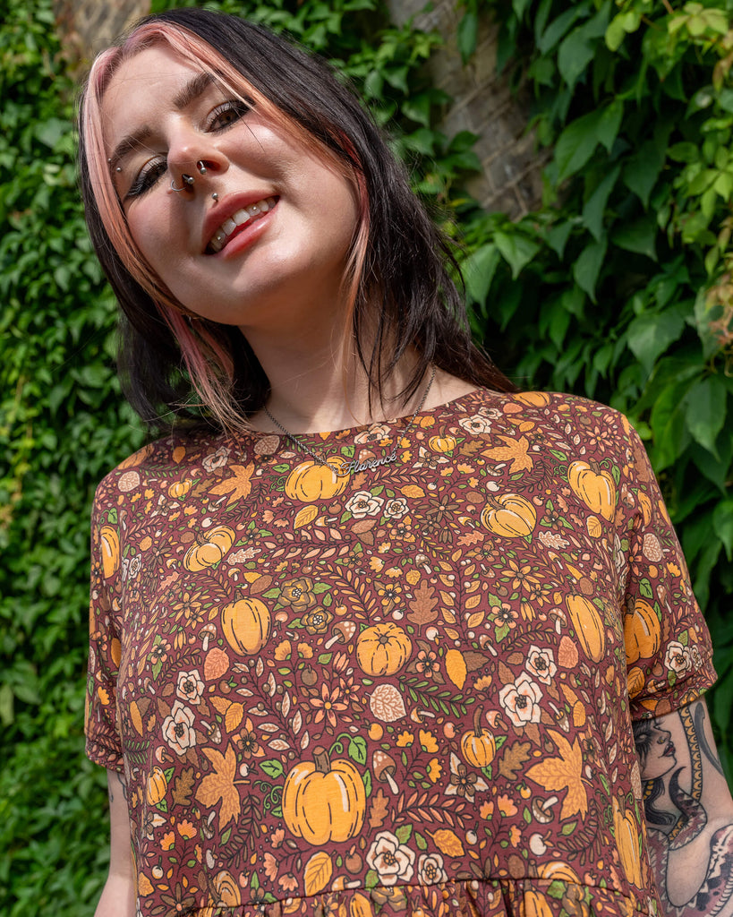 A smiling person wearing a Run & Fly smock dress with an autumnal pumpkin print. The dress features warm earthy tones, and the person is accessorised with a necklace that says "Florence." Tattoos are visible on their arms, with a background of green foliage. The brand is a small ethical slow fashion company.