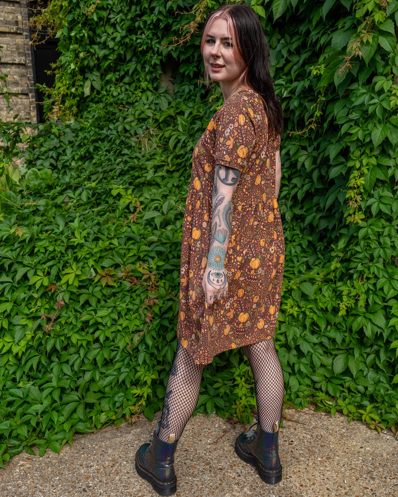 A person with tattoos is wearing a Run & Fly smock dress with an autumn-inspired print featuring pumpkins, mushrooms, and leaves. The person is standing outdoors, in front of a lush wall of greenery. The brand is a small ethical slow fashion company.