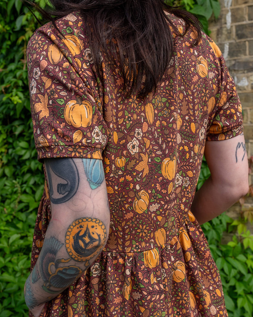 Close-up of the back of a Run & Fly smock dress with an autumn pumpkin print. The design includes pumpkins, leaves, and mushrooms, all in warm tones of orange and brown. The person has visible arm tattoos and is standing against a backdrop of greenery. The brand is a small ethical slow fashion company.