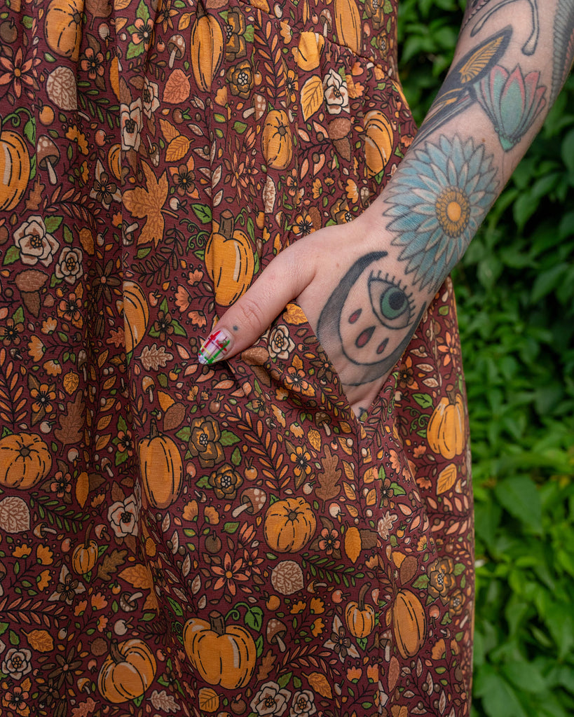 A close-up of a hand in the pocket of a Run & Fly smock dress. The fabric has an autumnal design of pumpkins, leaves, mushrooms, and acorns, all in warm earthy tones. Tattoos decorate the person's arm, and the background is lush greenery. The brand is a small ethical slow fashion company.
