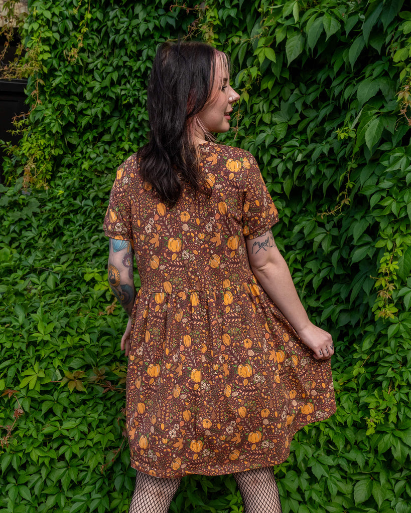 A person with tattoos stands with their back to the camera, wearing a brown smock dress by Run & Fly. The dress features a fun autumn print with orange pumpkins, acorns, mushrooms, and leaves. The person is in front of a background of vibrant green leaves. The brand is a small ethical slow fashion company.