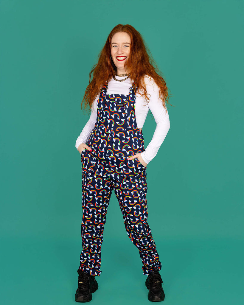 Isobella a white femme model with long ginger hair is wearing navy blue dungarees with all over rainbow and cloud print paired with long sleeve white top and black trainers. Isobella is smiling at the camera with her hands in the pockets of the dungarees in front of a green background. 