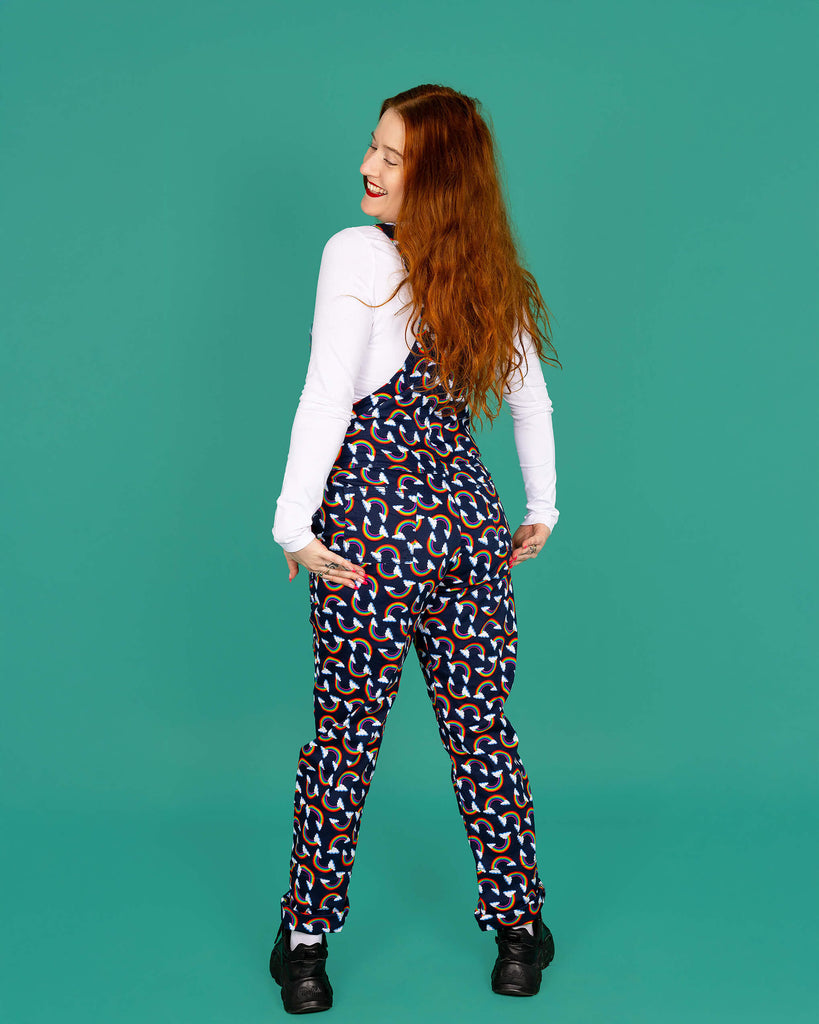 Isobella a white femme model with long ginger hair is wearing navy blue dungarees with all over rainbow and cloud print paired with long sleeve white top and black trainers. Isobella is stood with her back to the camera and is looking back over her shoulder smiling with her hands on the back of the dungarees. The background of the photo is green.