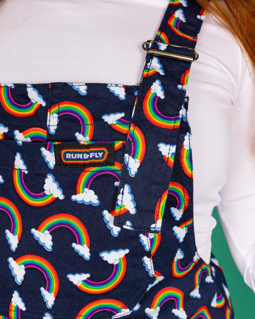 close up of Isobella wearing close up of navy blue dungarees with all over rainbow and cloud print paired with white top. Close up shows rainbow print in more detail and Run & Fly logo on the front pocket of the dungarees.