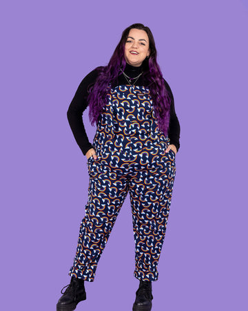 Luisa, a white femme model with purple hair, is wearing Over the Rainbow & Clouds Stretch Twill Dungarees with a long sleeve black turtle neck underneath and black boots. Luisa is stood in front of a lilac background on a black fluffy rug amongst disco balls, a star shaped light and a plant and is facing the camera smiling with her hands in the dungaree pockets.