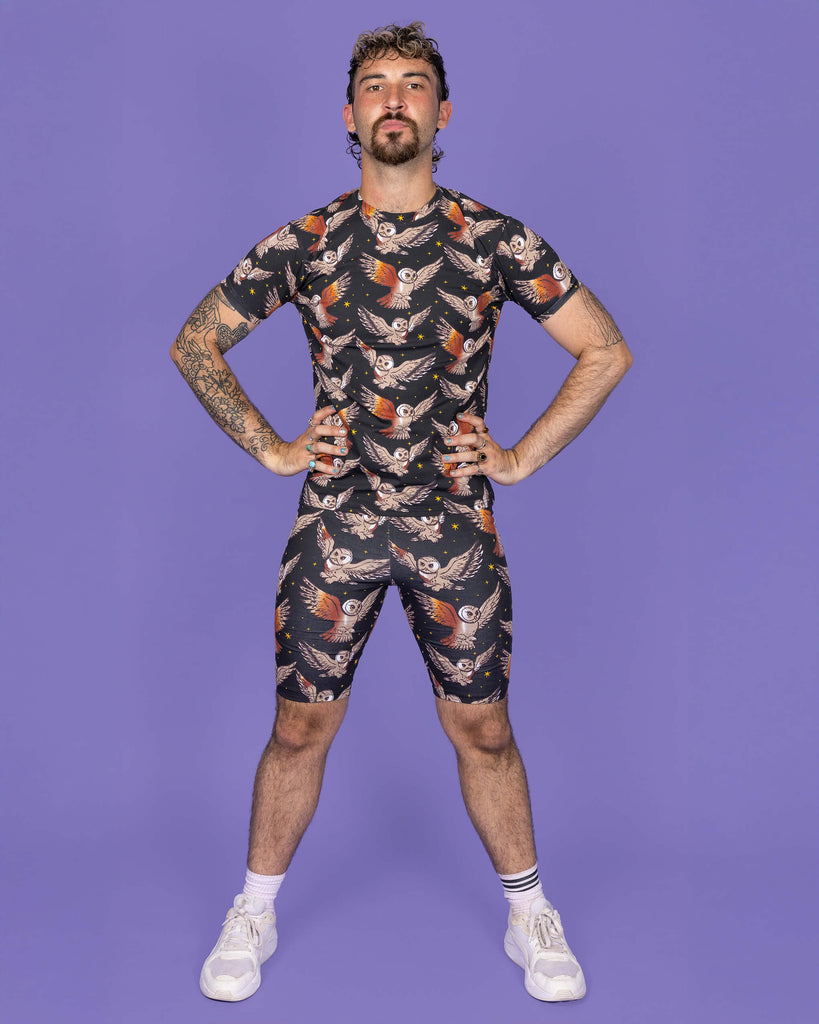 The What a Hoot Owl Stretch Cycle Shorts worn by a non binary masc person with a short curly brown mullet wearing the matching tshirt and white trainers on a purple studio background. They are facing forward pouting with their hands on their hips. The black base shorts features brown tawny owls flying amongst yellow stars.