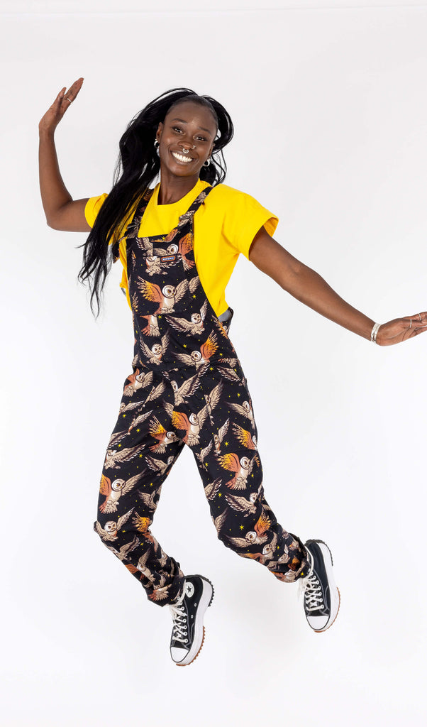 Yolande, a black femme model with a long black ponytail, is in a photography studio in Hove in front of a white backdrop wearing What A Hoot Owl Stretch Twill Dungarees with a bright yellow t shirt underneath and black trainers. The black dungarees have an all over print of cute barn owls and yellow stars. Yolande is jumping in the air with her arms up and is smiling at the camera.