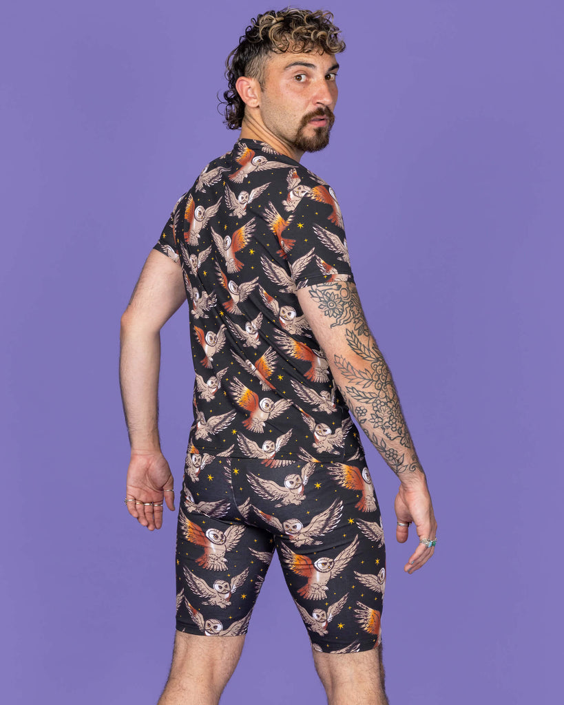 The What a Hoot Owl Stretch Cycle Shorts worn by a non binary masc person with a short curly brown mullet wearing the matching tshirt and white trainers on a purple studio background. They are facing away looking over their right shoulder to camera. The black base shorts features brown tawny owls flying amongst yellow stars.