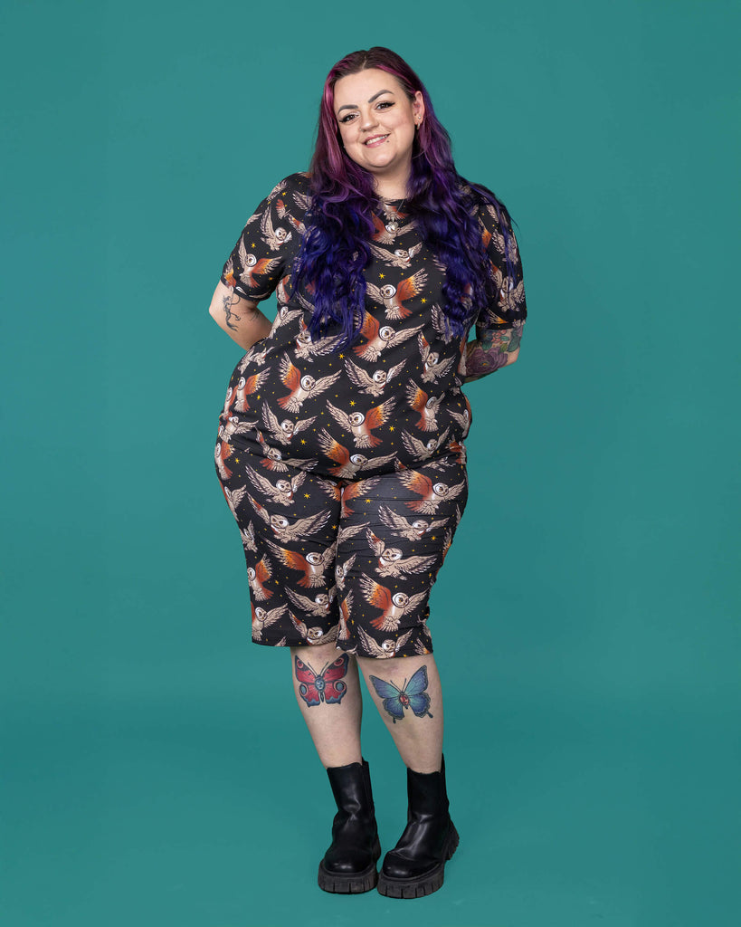 The What a Hoot Owl Stretch Cycle Shorts worn by a femme alternative tattooed model with long pink and purple hair, the matching tshirt and black boots on a teal studio background. She is facing forward smiling leaning forward with her hands resting behind her back. The black base shorts features brown tawny owls flying amongst yellow stars.