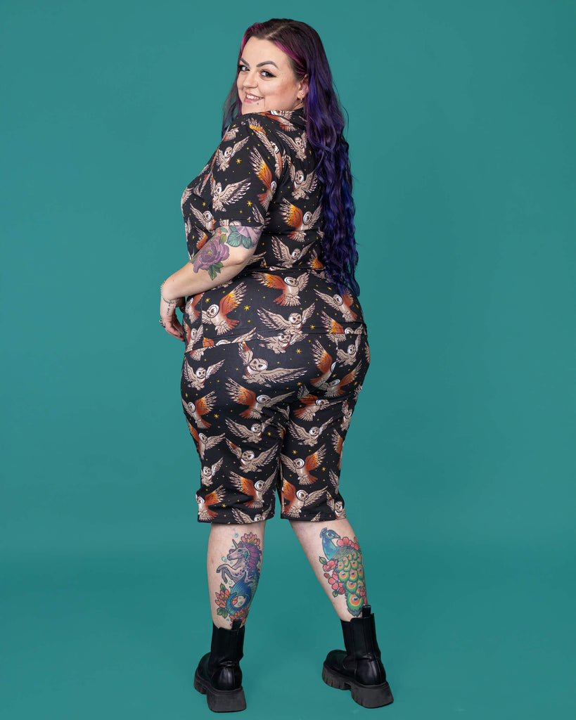 The What a Hoot Owl Stretch Cycle Shorts worn by a femme alternative tattooed model with long pink and purple hair, the matching tshirt and black boots on a teal studio background. She is facing away from camera smiling looking over her left shoulder with both arms resting on her hips. The black base shorts features brown tawny owls flying amongst yellow stars.