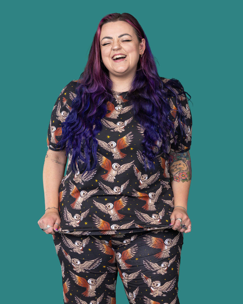 The What a Hoot Owl Short Sleeve T Shirt worn by a femme alternative tattooed model with long pink and purple hair, the matching cycle shorts on a teal studio background. She is facing forward laughing with her arms out to the side. The black base tshirt features brown tawny owls flying amongst yellow stars.