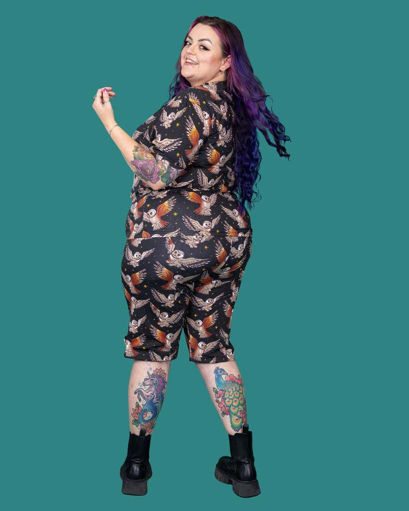 The What a Hoot Owl Short Sleeve T Shirt worn by a femme alternative tattooed model with long pink and purple hair, the matching cycle shorts and black boots on a teal studio background. She is facing away smiling looking over her left shoulder flicking her hair. The black base tshirt features brown tawny owls flying amongst yellow stars.