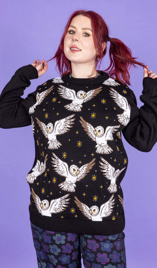 model with red hair wearing a black knit jumper with owls and stars on, paired with black jeans with blue and purple flowers on. The model is posing with her hands in her hair in front of a purple background.