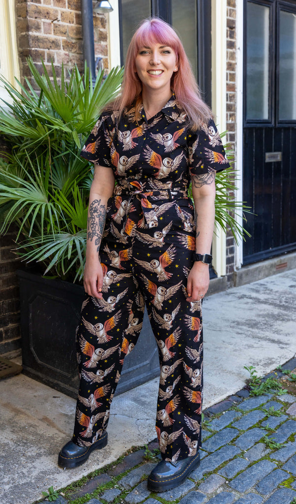 Stacie, a femme model with pink hair and tattoos, is outside in a mews in Hove on a sunny day wearing What a Hoot Owl Stretch Zip Jumpsuit with chunky black boots. The black jumpsuit has an all over print of cute barn owls and yellow stars. Stacie is stood facing the camera with ehr arms down by her sides.