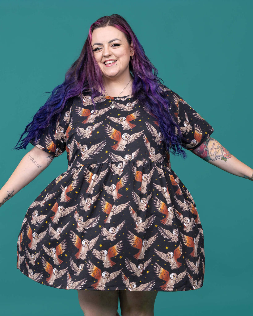The What a Hoot Owl Oversized Smock Dress worn by a femme alternative tattooed model with long pink and purple hair, the matching tshirt and black boots on a teal studio background. She is facing forward smiling twirling in the dress with her arms out to the side. The black base dress features brown tawny owls flying amongst yellow stars.