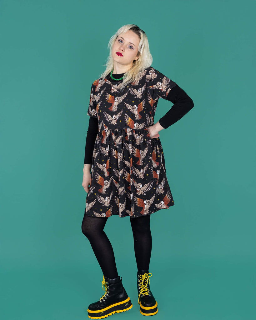 The What a Hoot Owl Oversized Smock Dress worn by a femme model with white blonde hair, a long black tshirt, black tights and yellow black boots on a teal studio background. She is facing forward leaning back on one leg whilst the other is bent forward, she has one hand on her hip and is leaning her head back smising to camera. The black base dress features brown tawny owls flying amongst yellow stars.